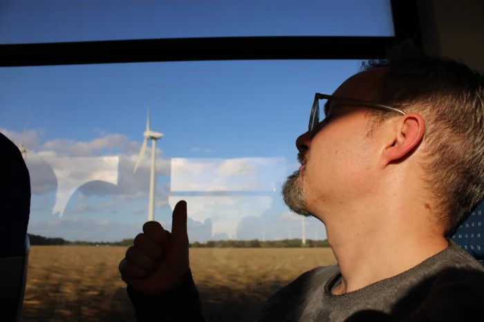 Ein Mann mit Brille sitzt im Zug, draußen sind Felder und Windräder zu sehen.
